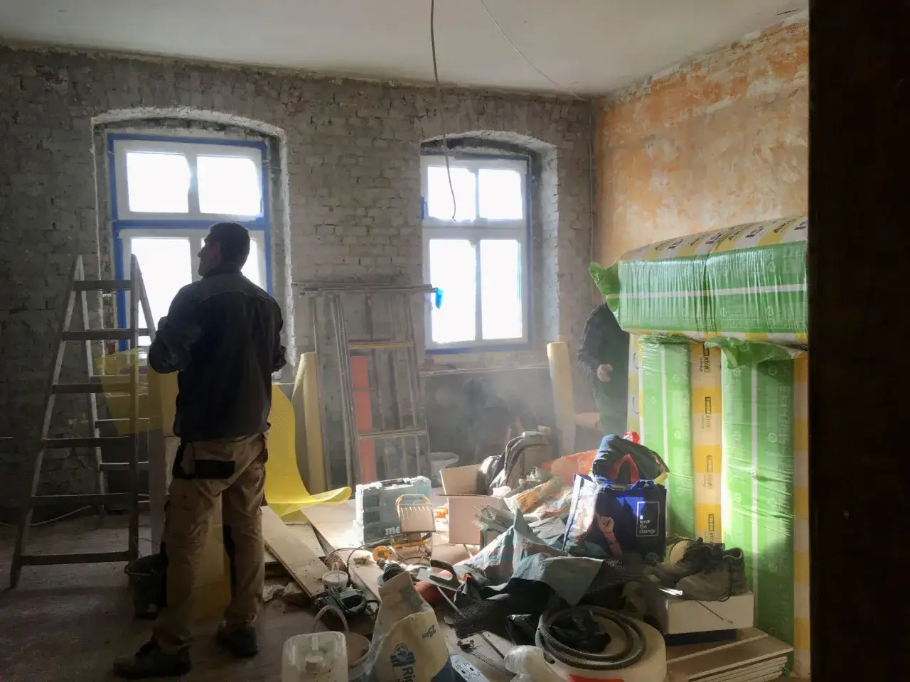 Remontowany pokój z dwoma oknami, drabina, materiały budowlane i pracownicy.